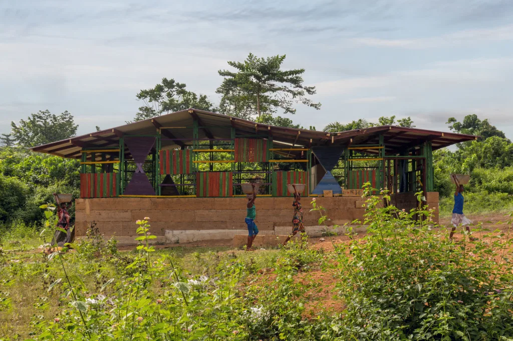 Personas caminan frente a una estructura rural construida con bloques de tierra y paneles de colores, rodeada de vegetación.