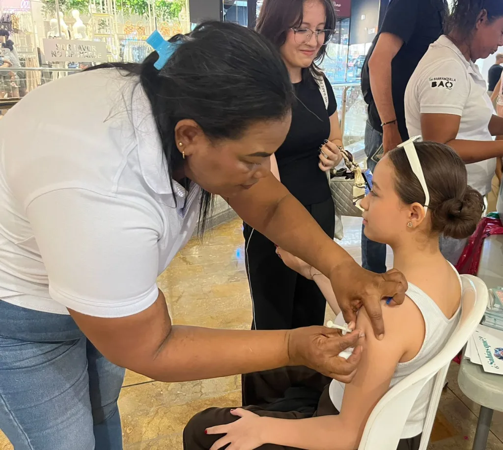 Trabajadora de la salud aplicando una vacuna en el brazo de una niña sentada en una silla plástica, en un espacio público con personas alrededor y vitrinas comerciales al fondo.