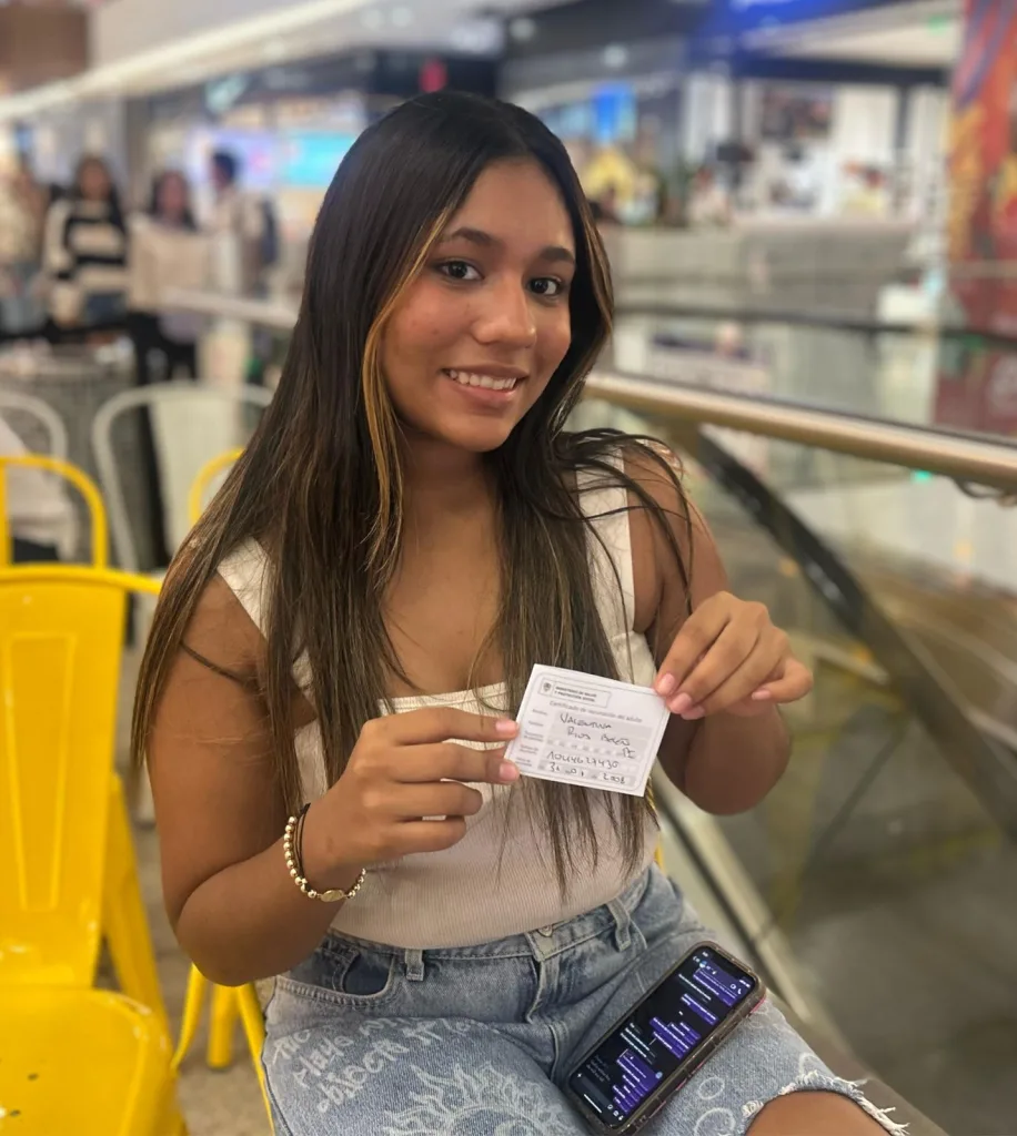Mujer joven sentada en un centro comercial sosteniendo una tarjeta de vacunación con su primera dosis, sonriendo a la cámara.