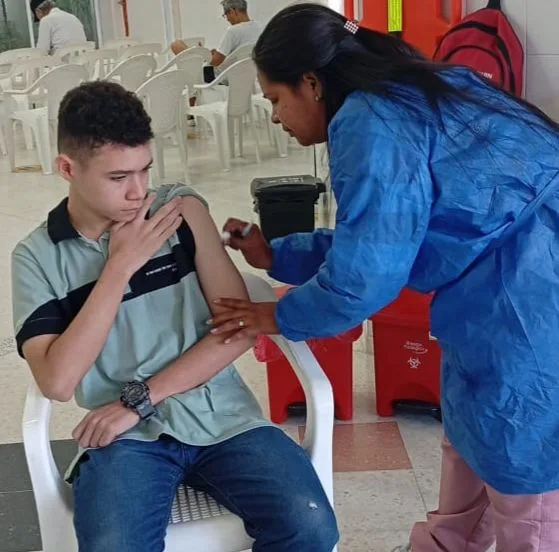Persona joven recibiendo una vacuna en el brazo por parte de una trabajadora de la salud con bata azul, en un punto de vacunación con sillas plásticas y contenedores de residuos biosanitarios al fondo.