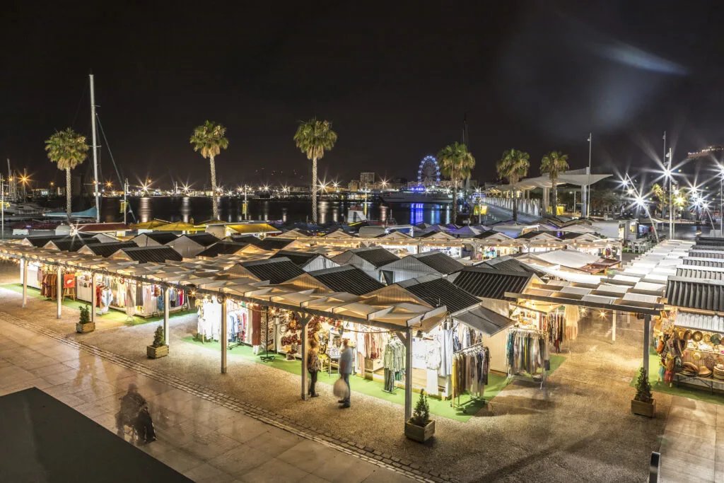 Mercadillo nocturno en el puerto de Málaga con puestos iluminados, palmeras y noria al fondo.