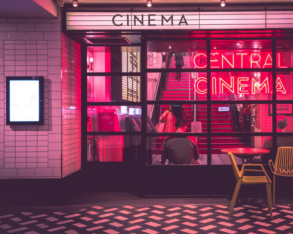 Entrada iluminada de un cine con luces de neón rosadas y rojas, con personas en el interior