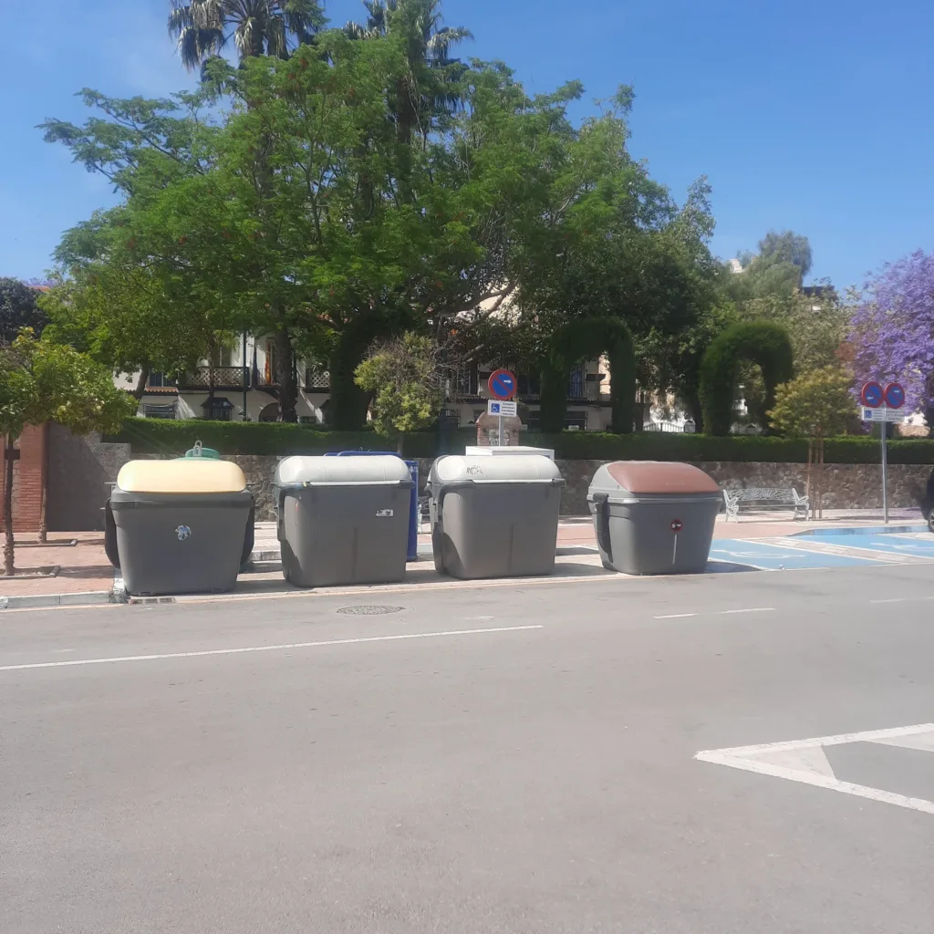 Fila de contenedores de reciclaje y residuos en una calle de Churriana, con árboles y viviendas alrededor.