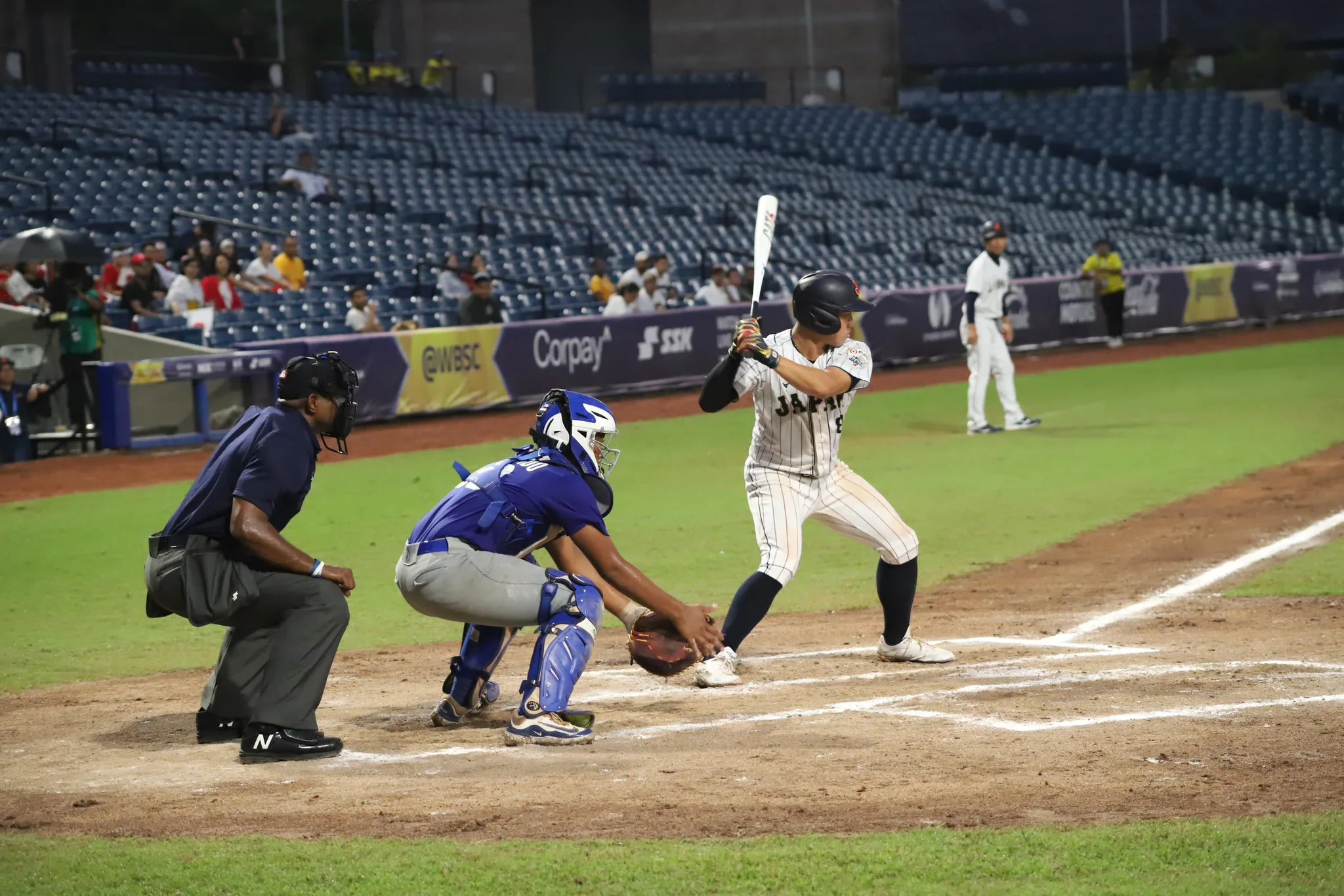 Bateador japonés en acción durante juego de béisbol con catcher y árbitro en estadio iluminado